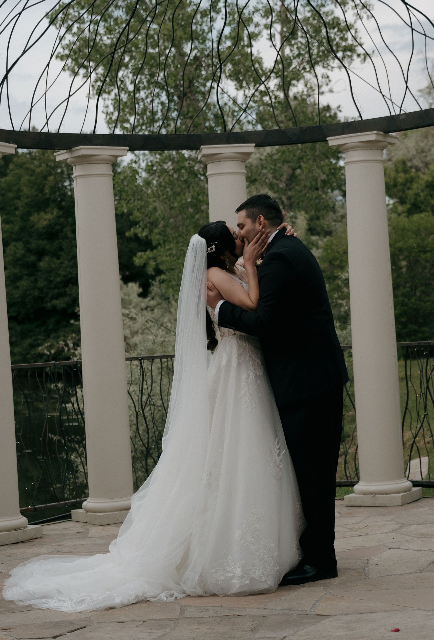Bride and groom share their first kiss at outdoor wedding altar surrounded by elegant white columns and lush greenery.