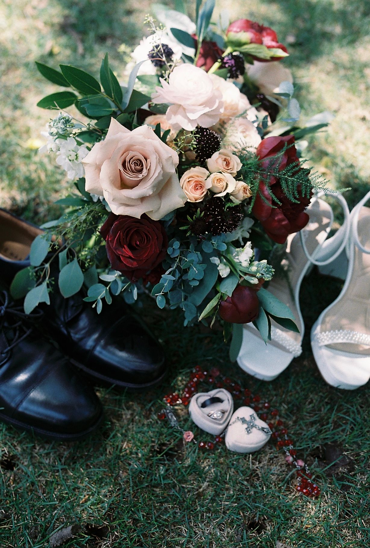 Wedding bouquet with blush and burgundy roses surrounded by black dress shoes and white bridal heels on green grass.
