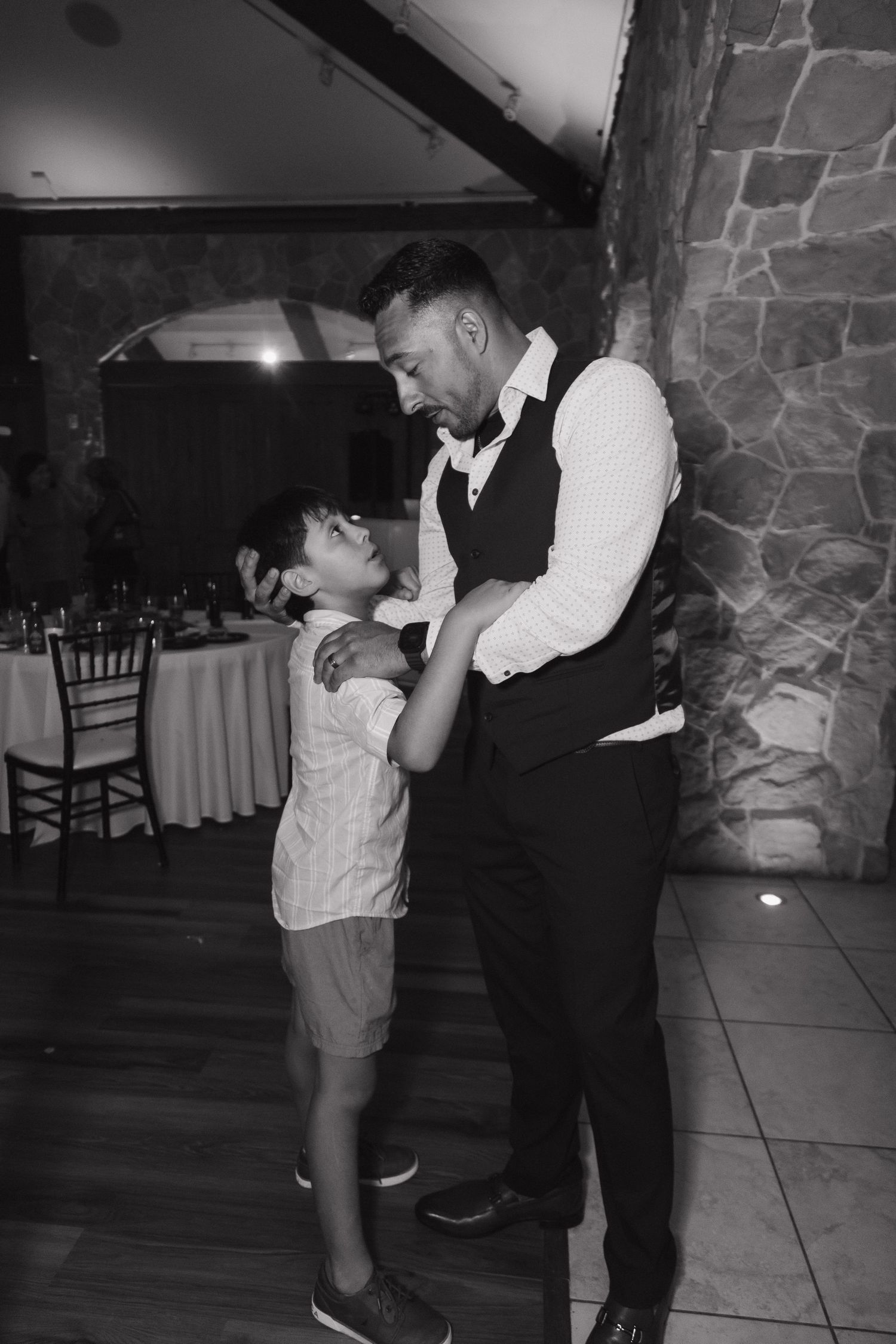 Black and white photo of groom in vest sharing a tender moment dancing with young boy.