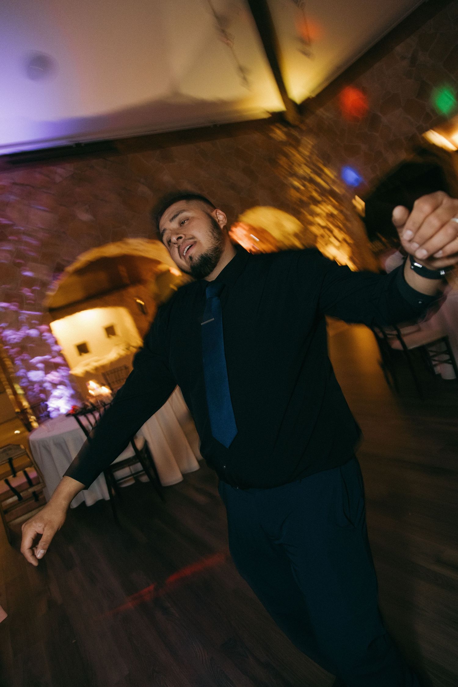 An energetic wedding guest in a black suit and blue tie dances enthusiastically on the reception dance floor.