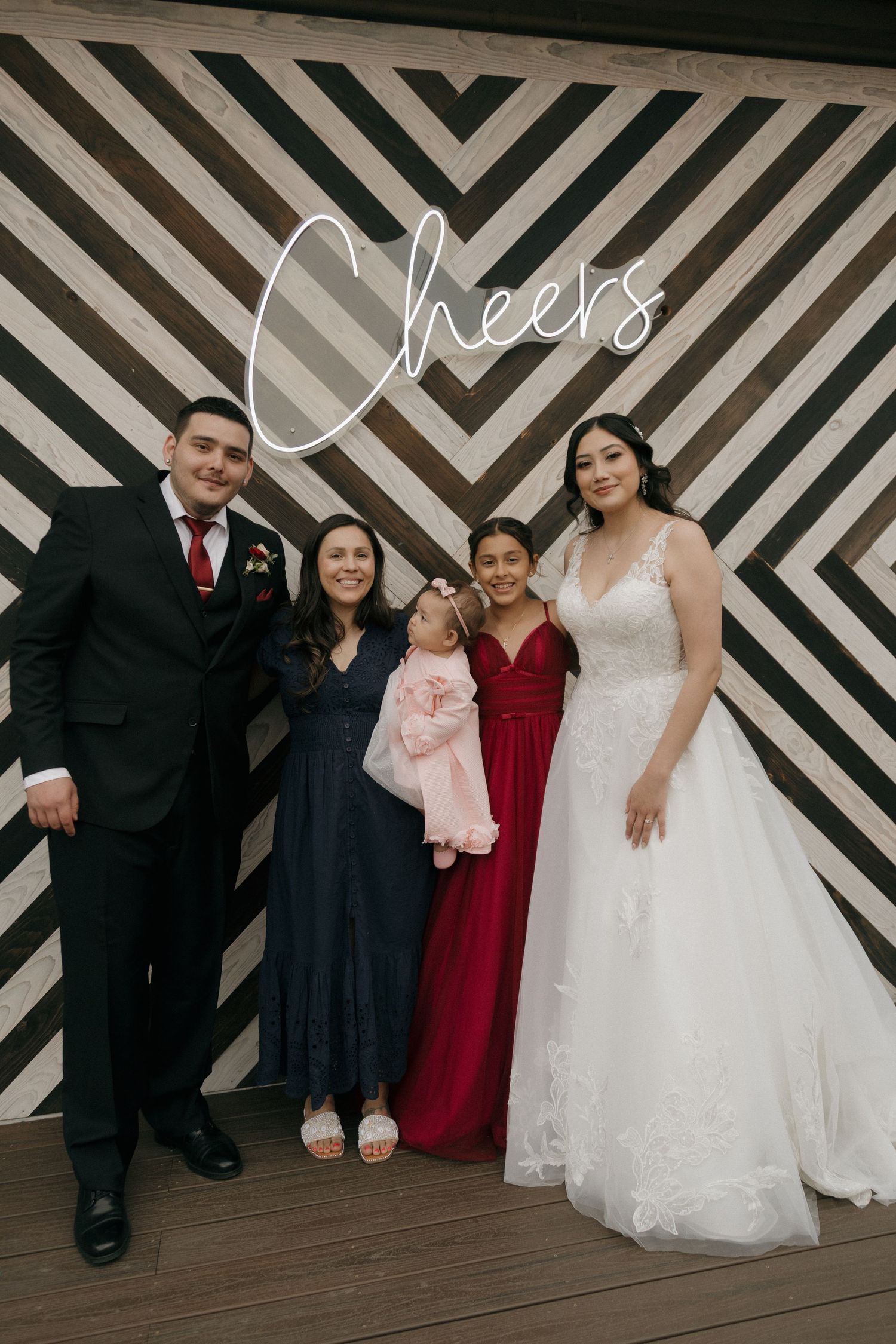 Bride in white lace gown and groom pose with family including children in front of Cheers neon sign backdrop.