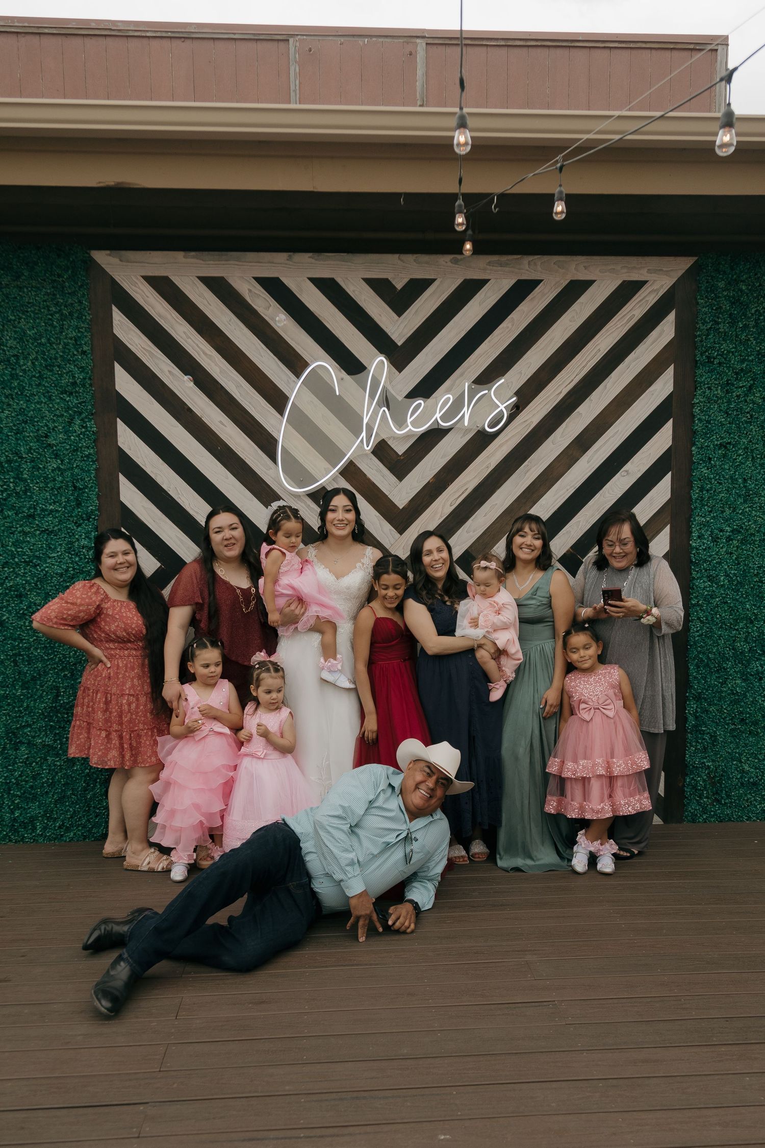 Bride in white gown poses with family group in front of Cheers neon sign.