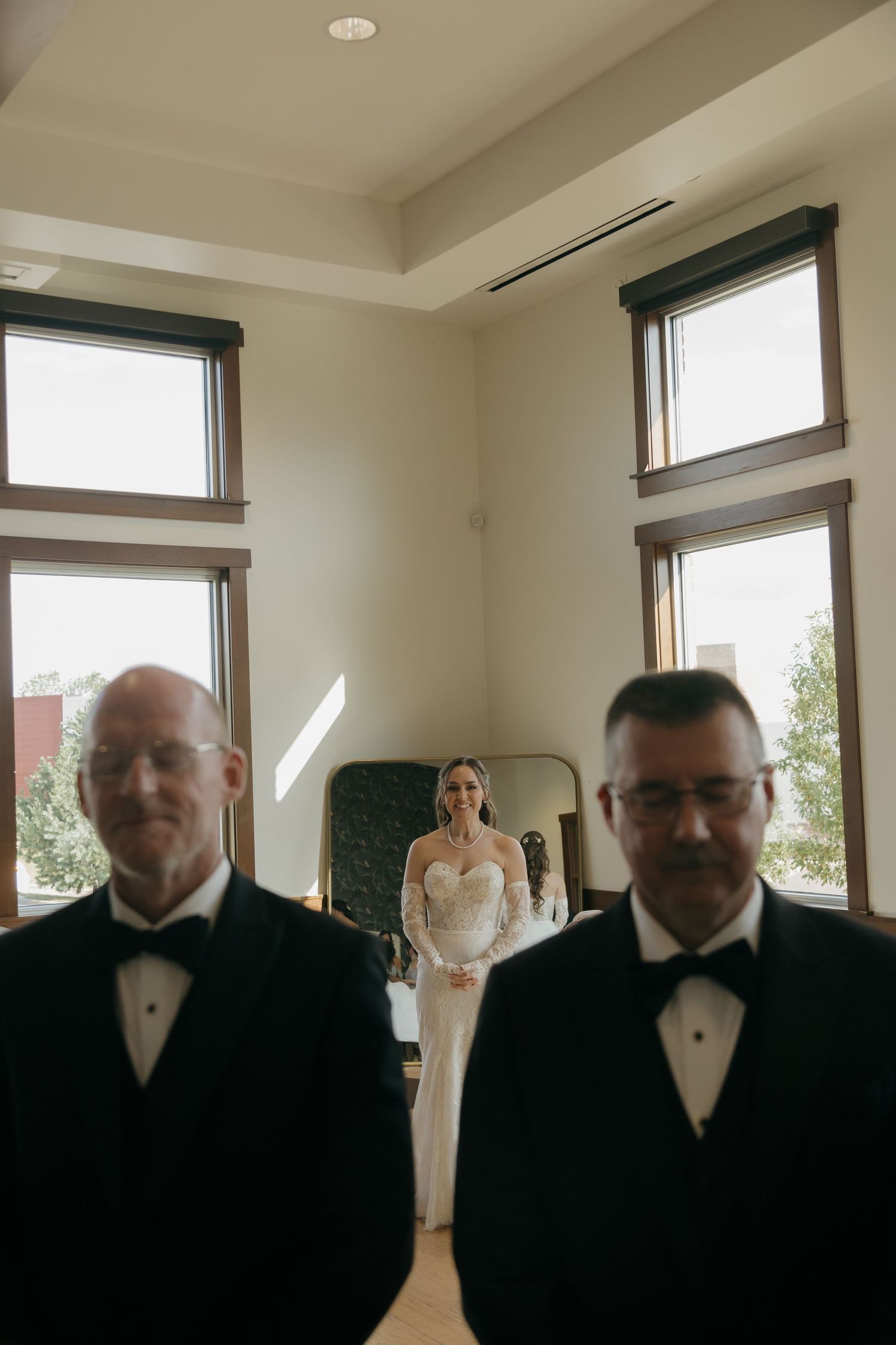 Bride in white gown stands behind two men in tuxedos during wedding first look moment indoors.
