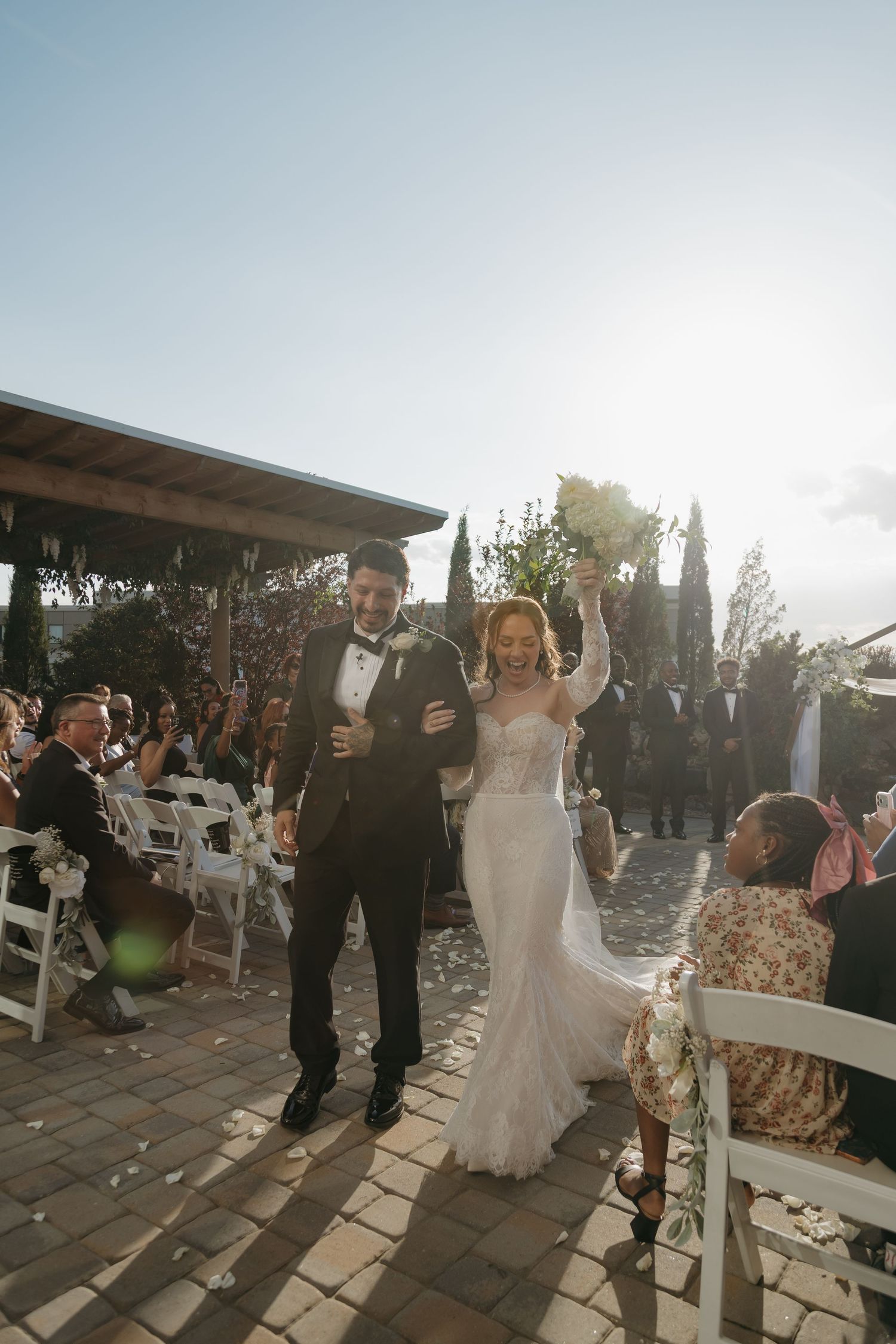 Bride and groom walk joyfully down the aisle after wedding ceremony outdoors in sunlight.
