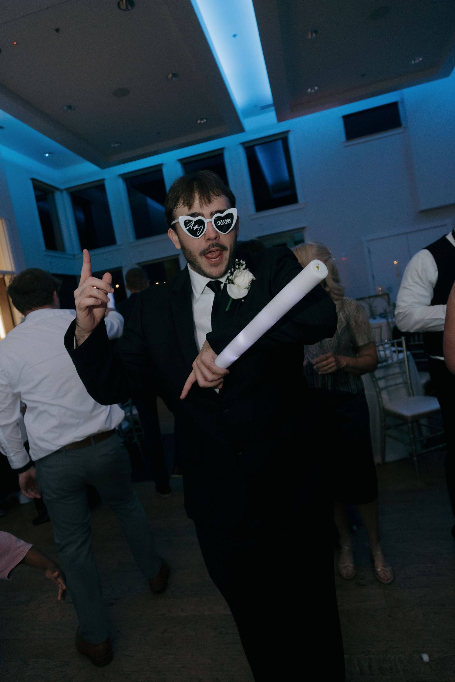 Wedding guest in black suit and heart sunglasses holding glow stick on dance floor.