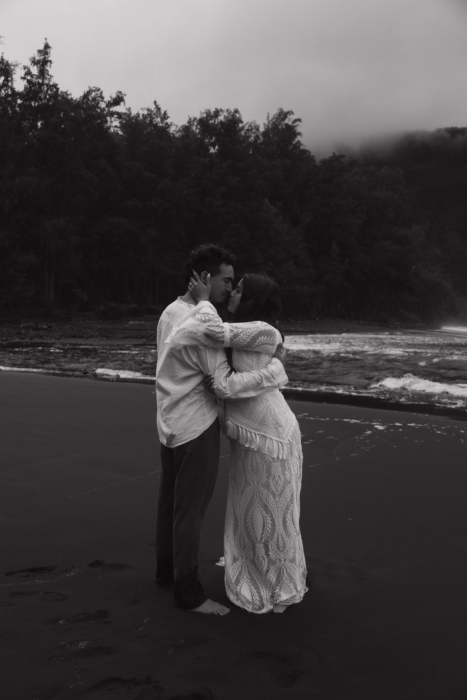 Black and white photo of newlyweds sharing a kiss on a dramatic black sand beach at dusk.