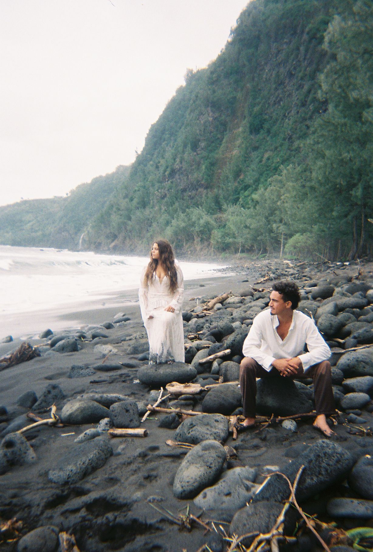 Couple on rocky black sand beach with lush green cliffs, woman in white dress standing, man sitting on rocks.