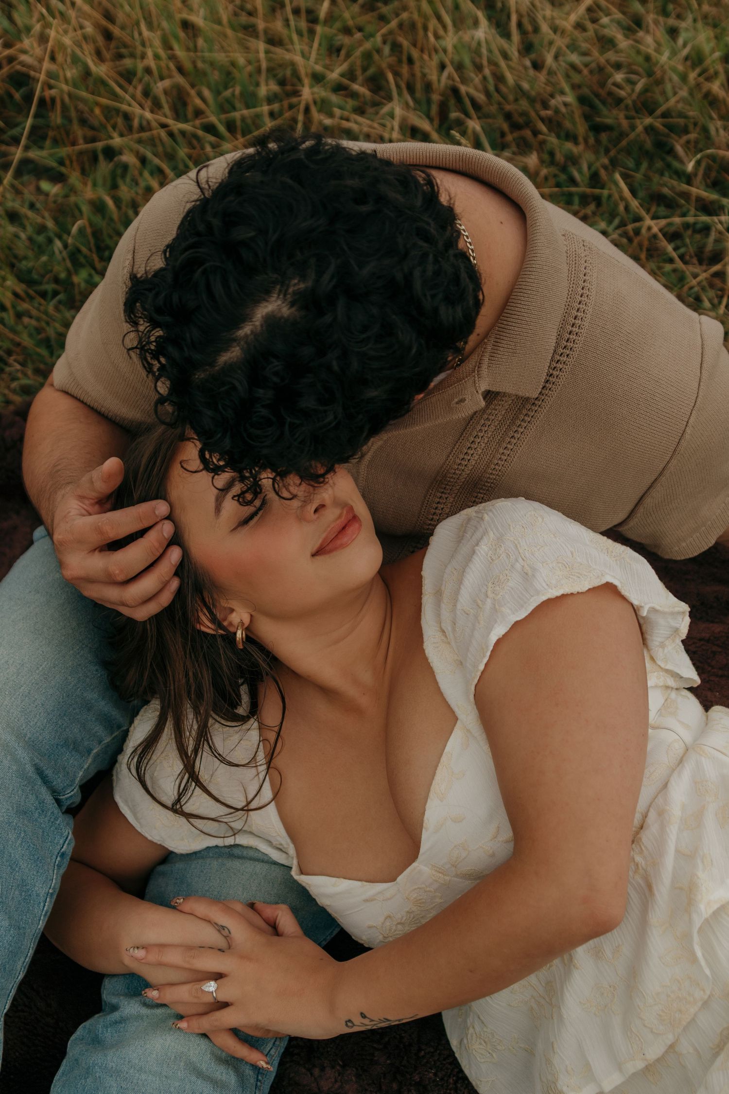 A couple shares an intimate moment lying in a grassy field, the man leaning down to kiss her.