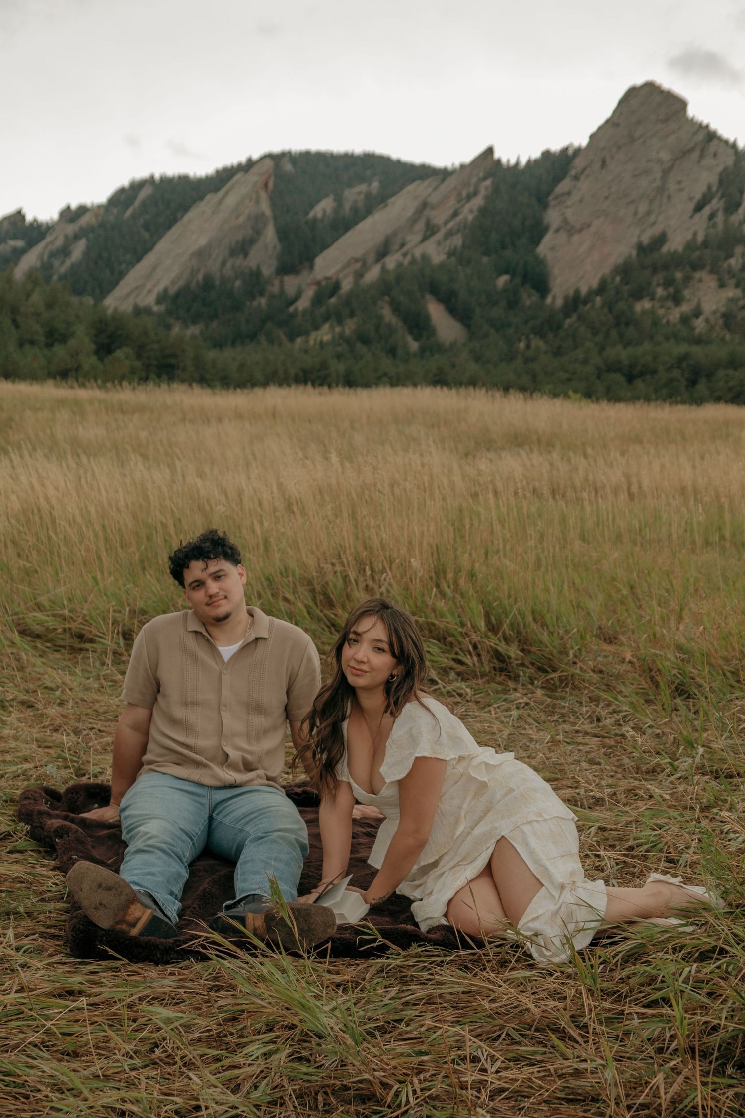 Couple sitting together in golden grass field with iconic Flatirons rock formations behind them.