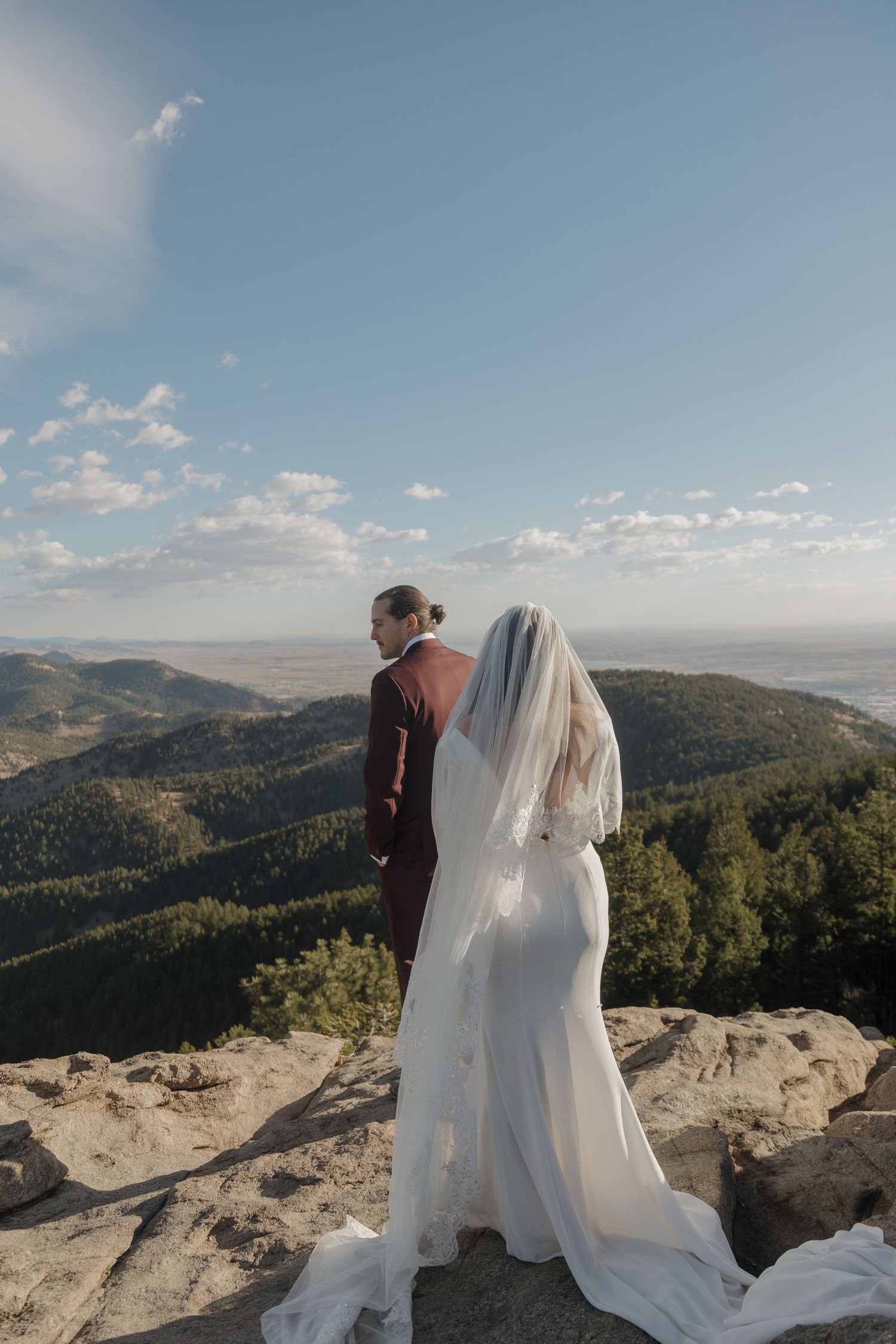 A bride and groom stand on a rocky mountain cliff overlooking a vast forested valley.
