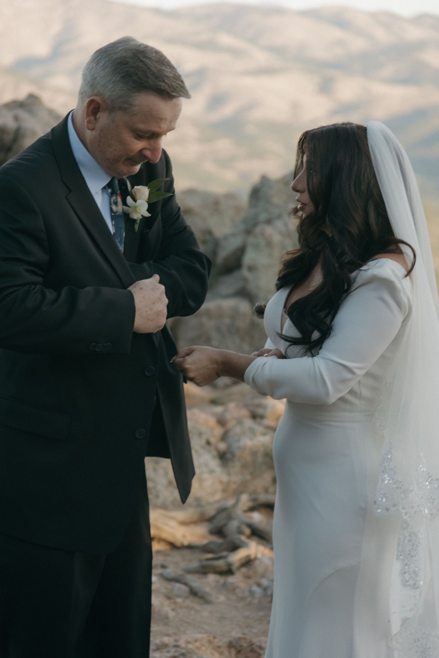 A bride places a wedding ring on the groom's finger during an outdoor mountain ceremony at dusk.