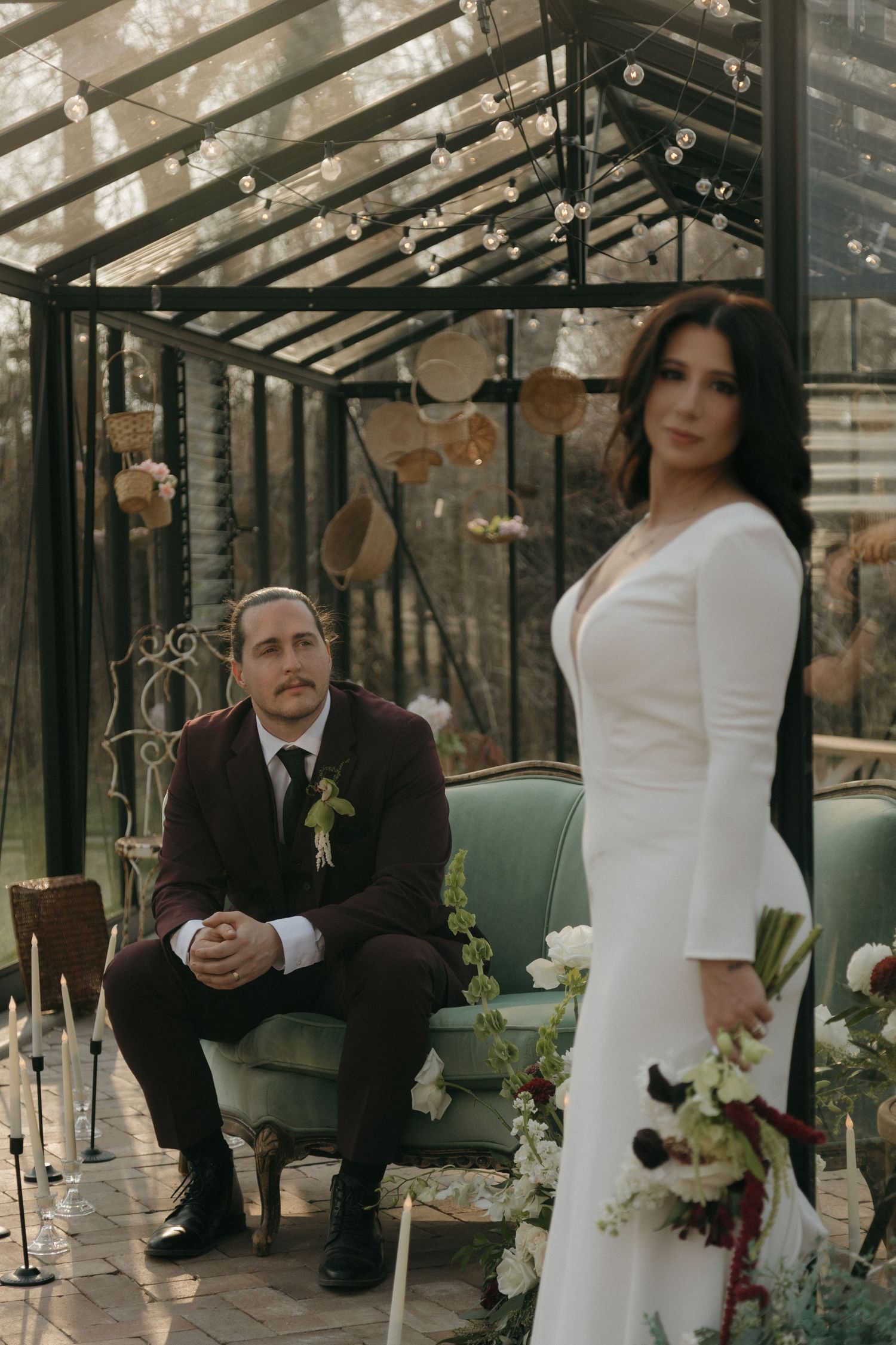 Bride in white gown and groom in burgundy suit posing in decorated glass greenhouse venue.