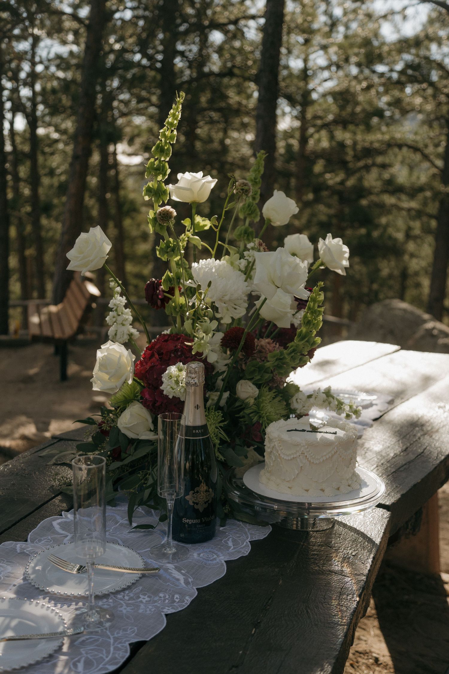 Elegant outdoor wedding table with white roses, red dahlias, champagne bottle, and white cake in forest setting.