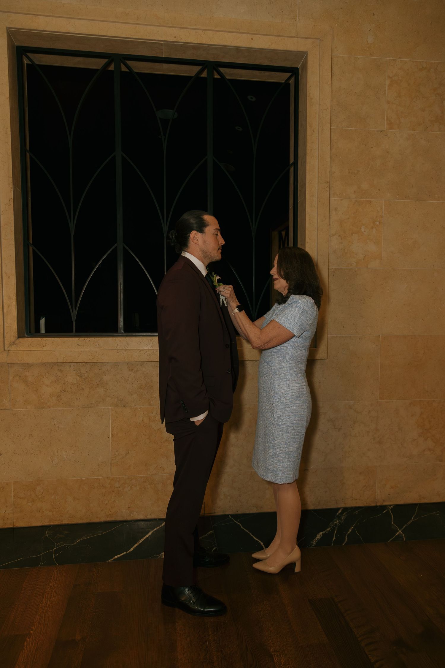 A woman in a blue dress pins a boutonniere on a man in a dark suit indoors.