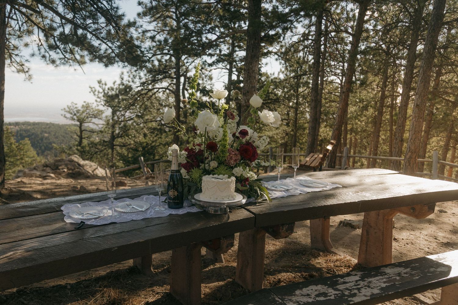 Rustic wooden table with wedding cake and floral arrangements set among pine trees outdoors.