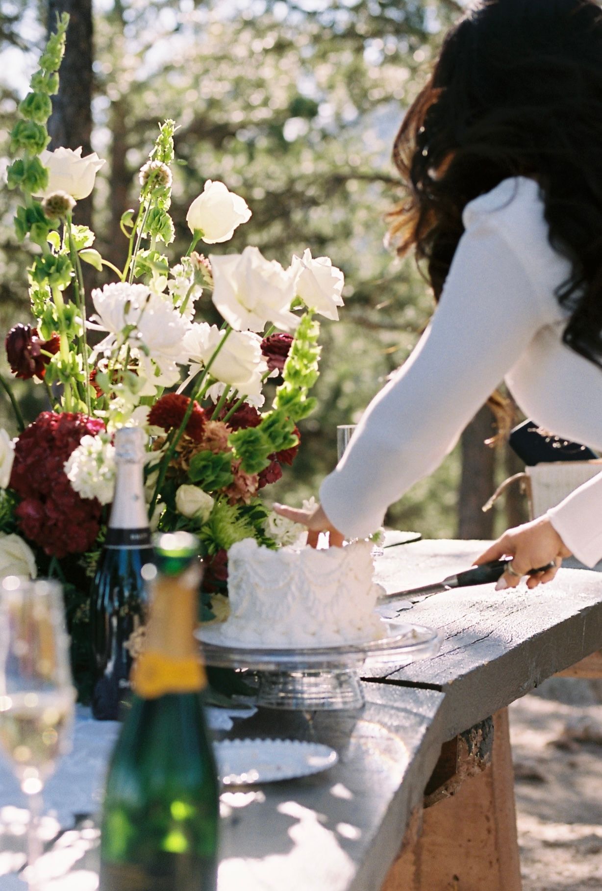 Bride in white long-sleeve gown cuts wedding cake beside lush floral arrangement outdoors.