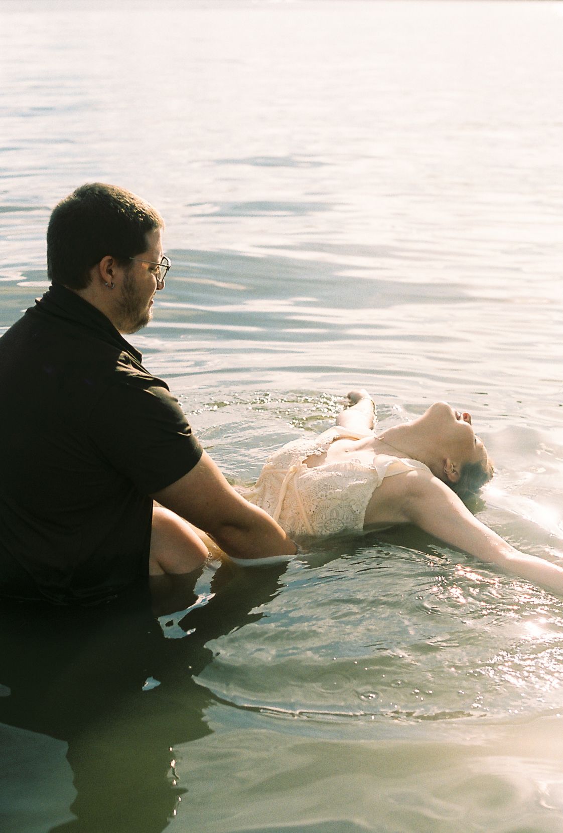 Man in black shirt supports woman floating in sunlit water during romantic outdoor photo session.