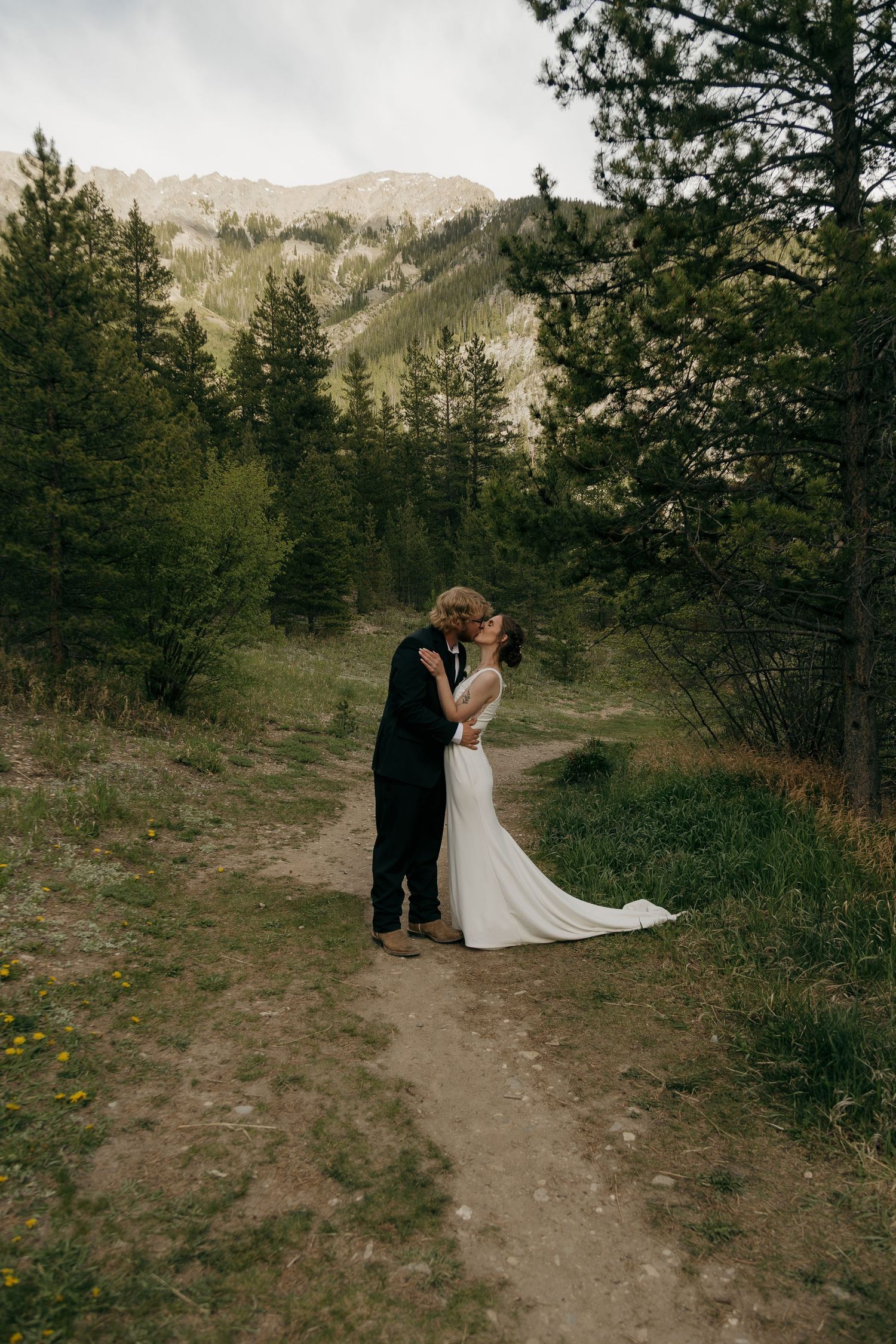 Bride and groom share a romantic kiss on a mountain trail surrounded by pine trees and scenic peaks.