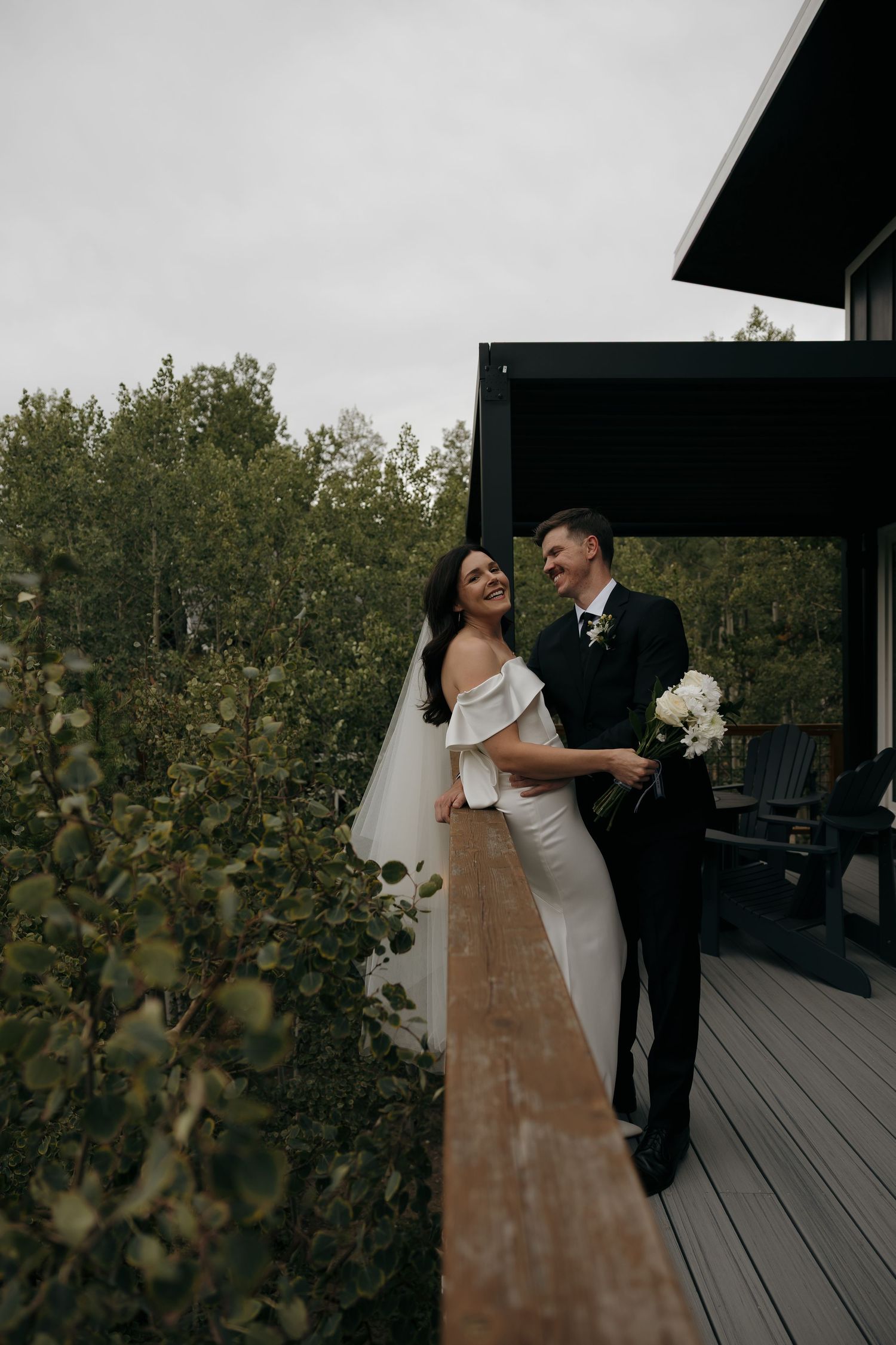 Laughing bride and groom embrace on wooden deck surrounded by lush green trees.