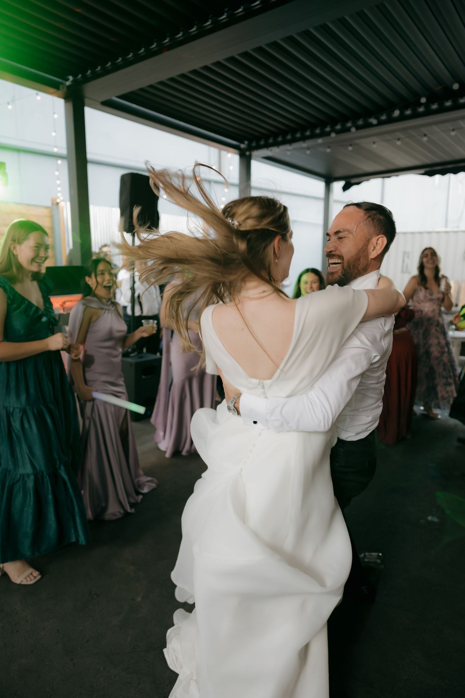 Groom laughs joyfully while spinning bride in white gown on wedding reception dance floor.