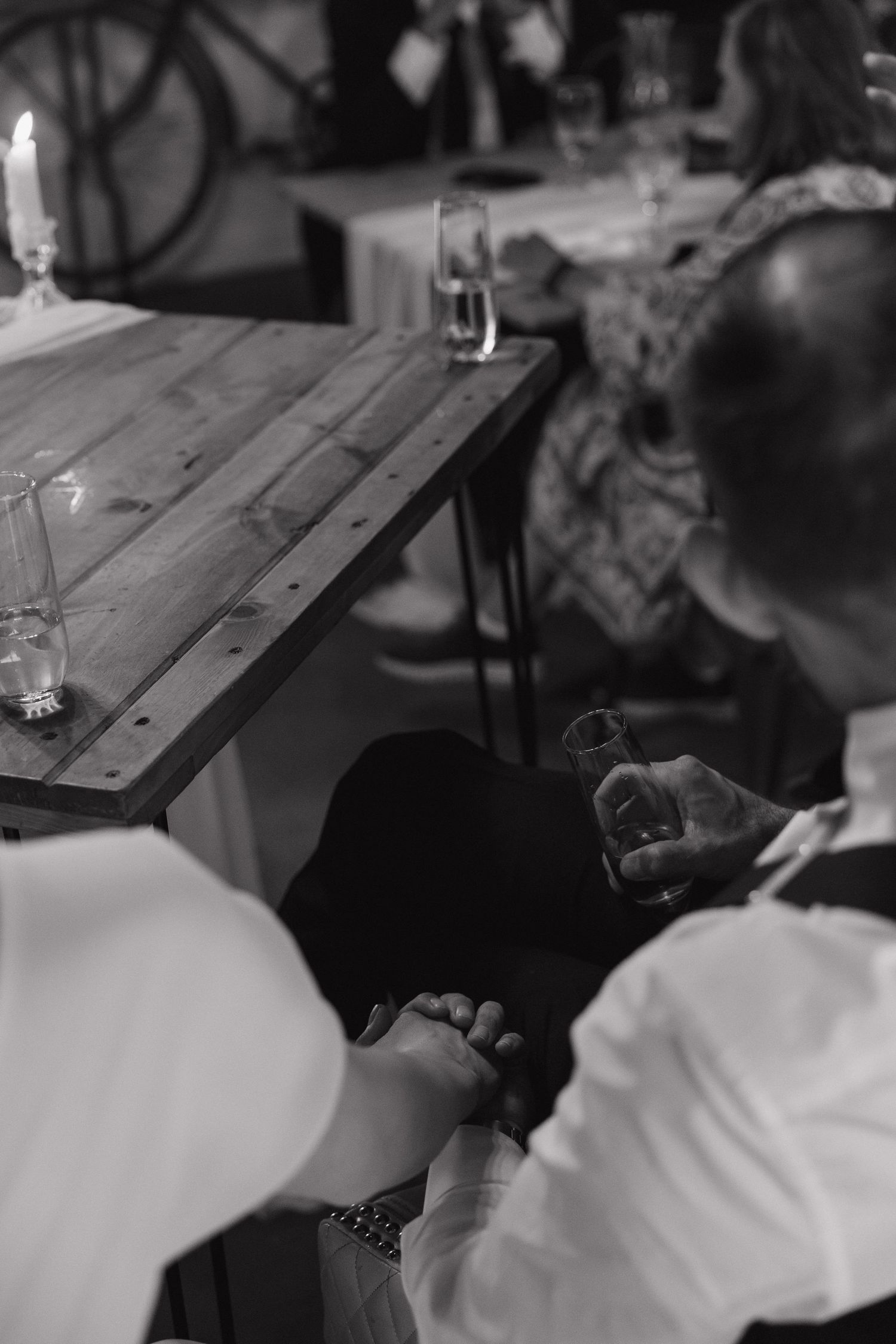 Black and white photo of couple holding hands at wedding reception table with candles and drinks.