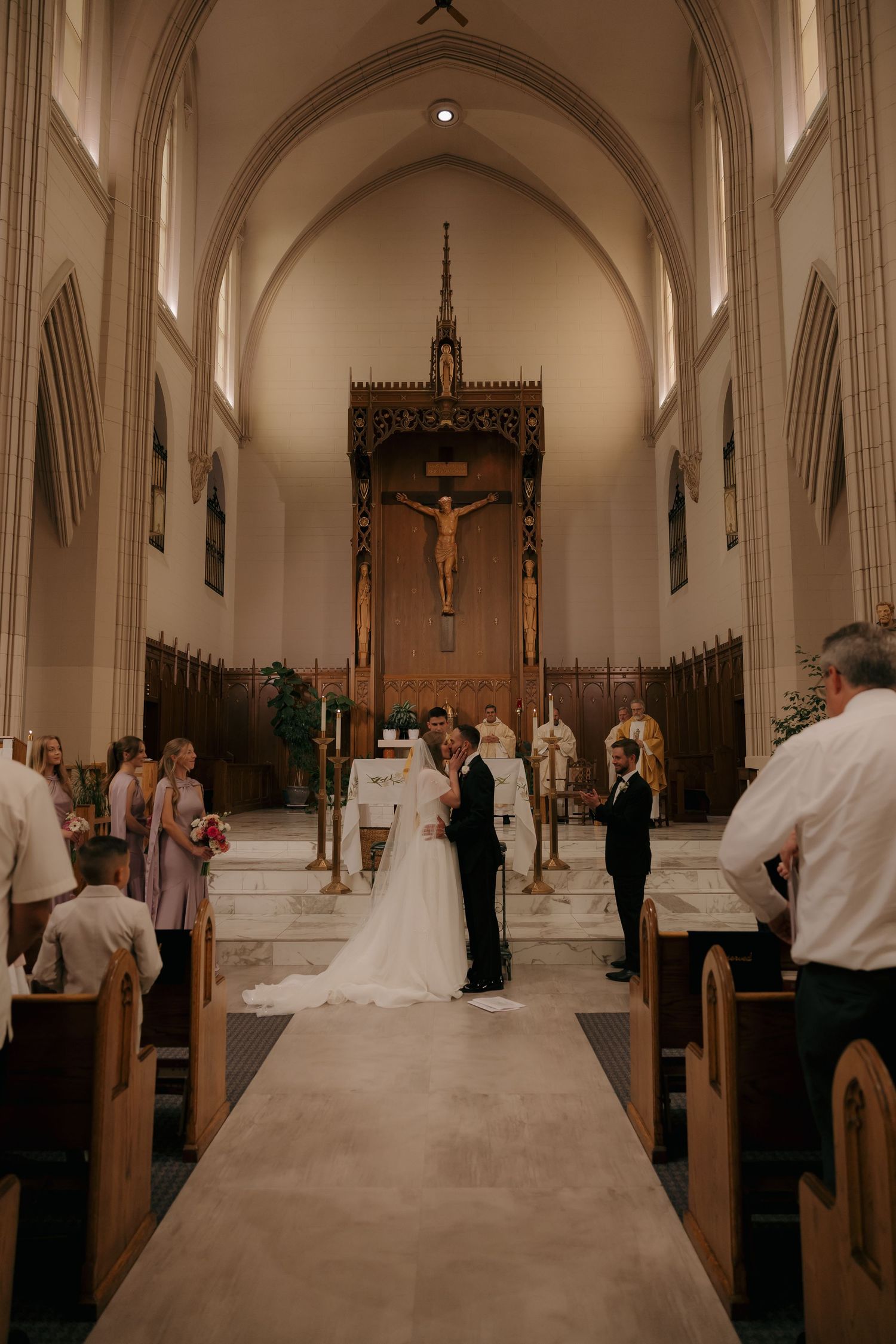 Bride and groom exchange vows at altar of elegant church with wooden crucifix and arched ceilings.