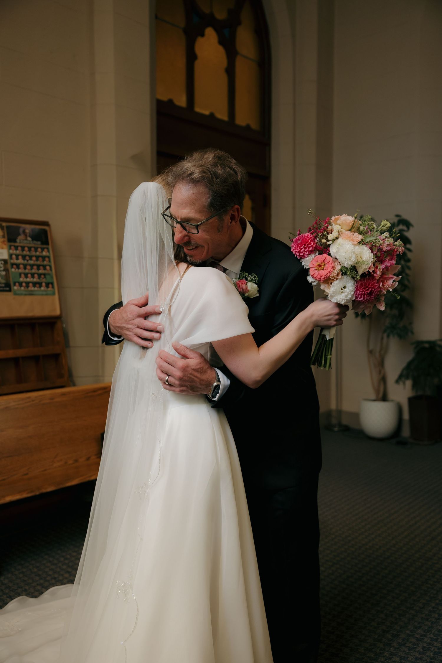 Emotional bride in white cape gown embraces older man, likely her father, while holding a pink and white floral bouquet.
