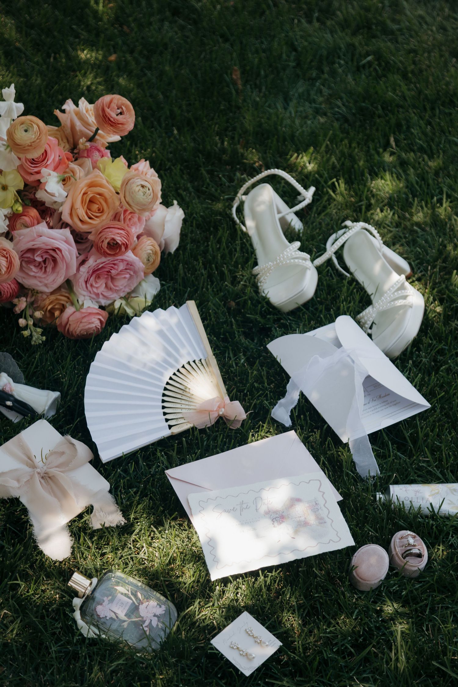 Wedding flat lay on green grass with pink roses bouquet, white heels, fan, envelope, perfume, and macarons.