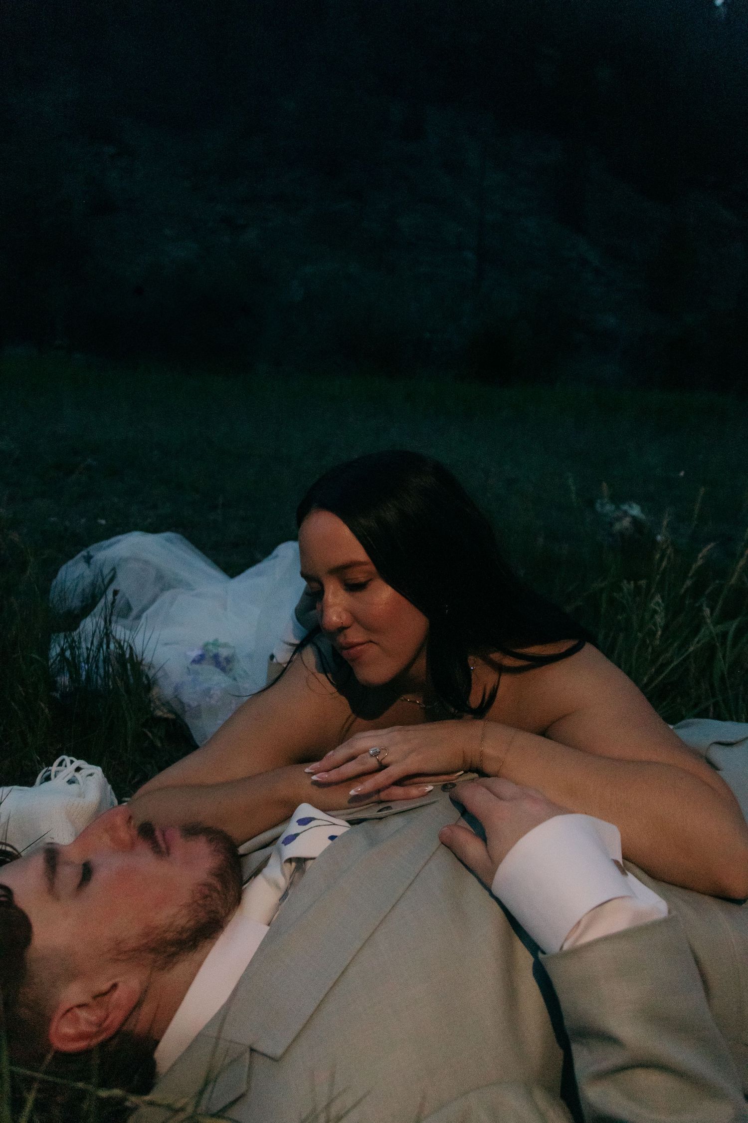 Bride and groom lying in grass at dusk in intimate romantic wedding portrait.