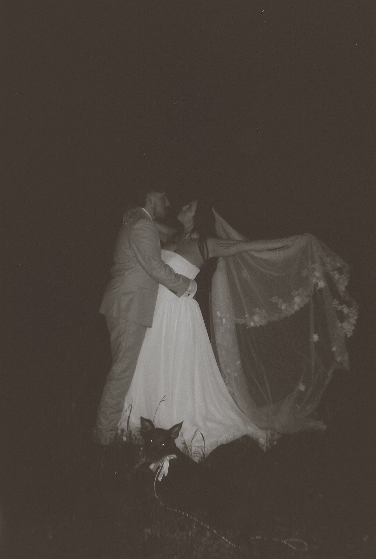 Black and white romantic photo of bride and groom kissing under billowing veil at night outdoors.