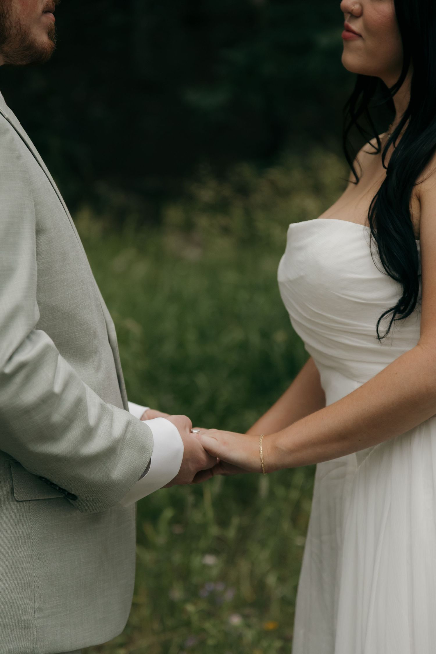 A bride in a white strapless gown and groom in a gray suit hold hands outdoors during a romantic wedding ceremony.