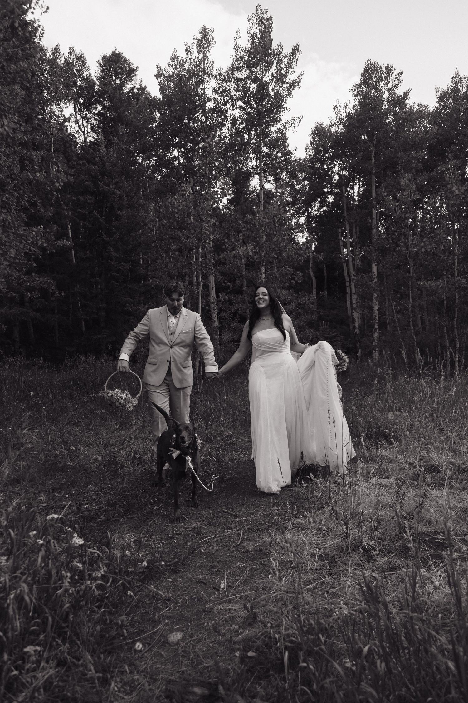 Black and white photo of bride and groom walking hand in hand with their dog through forest path.