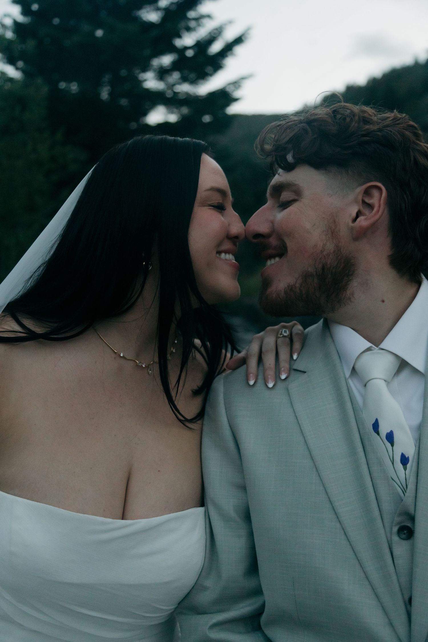 Close-up of smiling bride and bearded groom in gray suit sharing an intimate moment about to kiss outdoors.