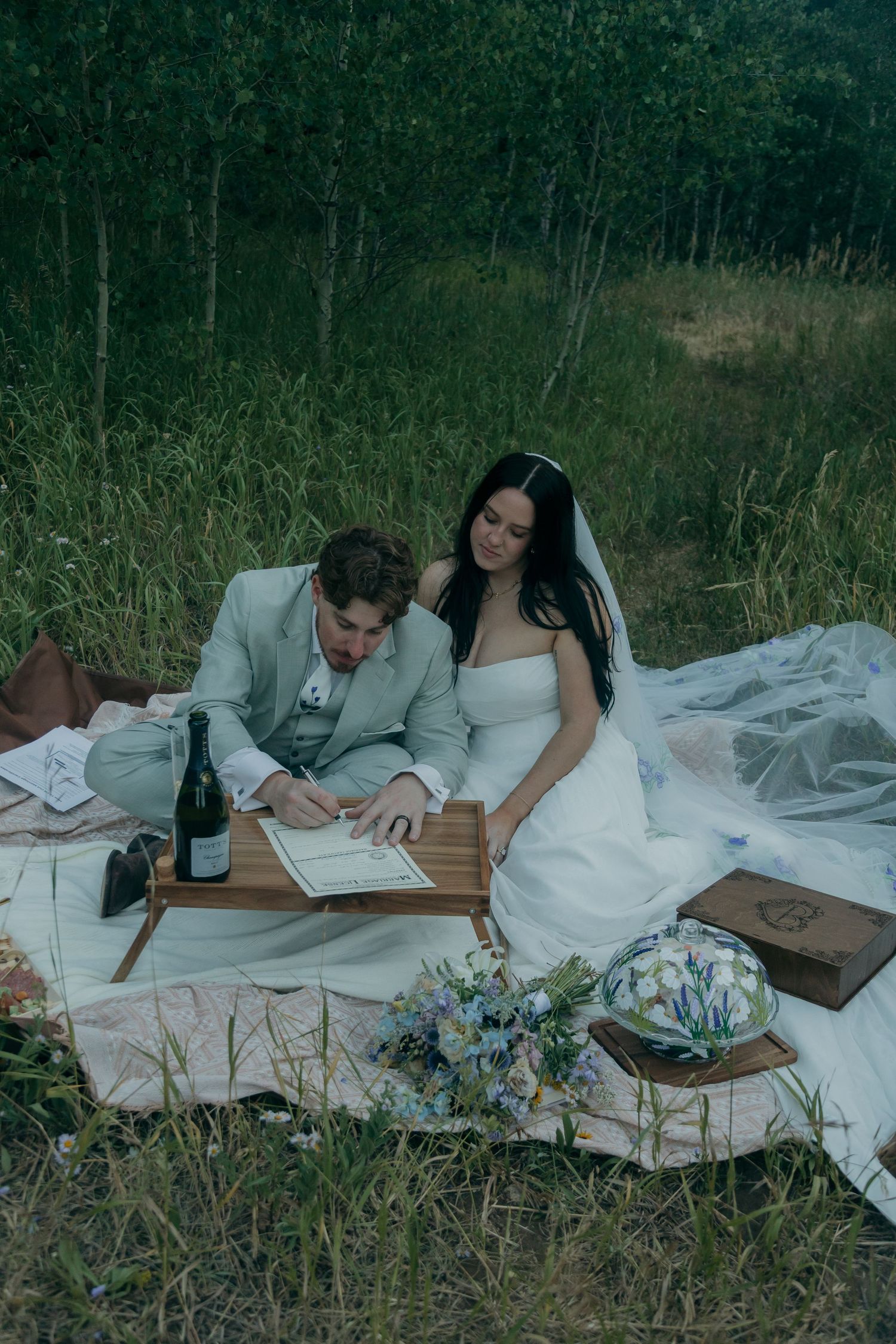 Bride and groom sign marriage license together on a picnic blanket in a lush green forest.