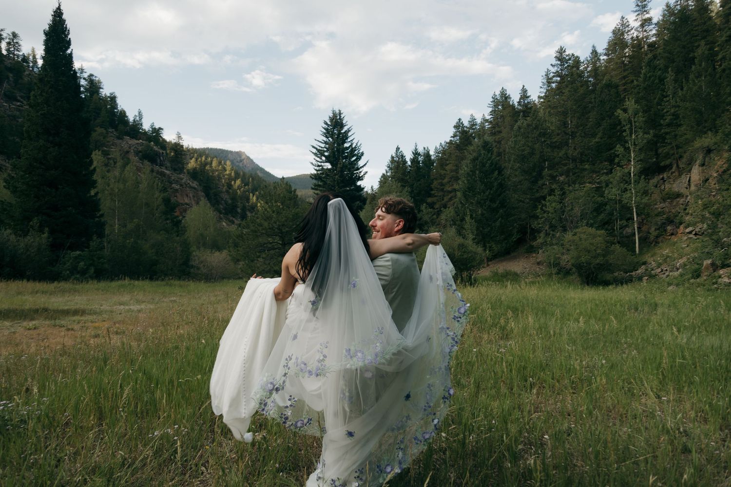 Groom lifts bride in a sweeping embrace in a green mountain meadow, her embroidered veil flowing dramatically in the breeze.