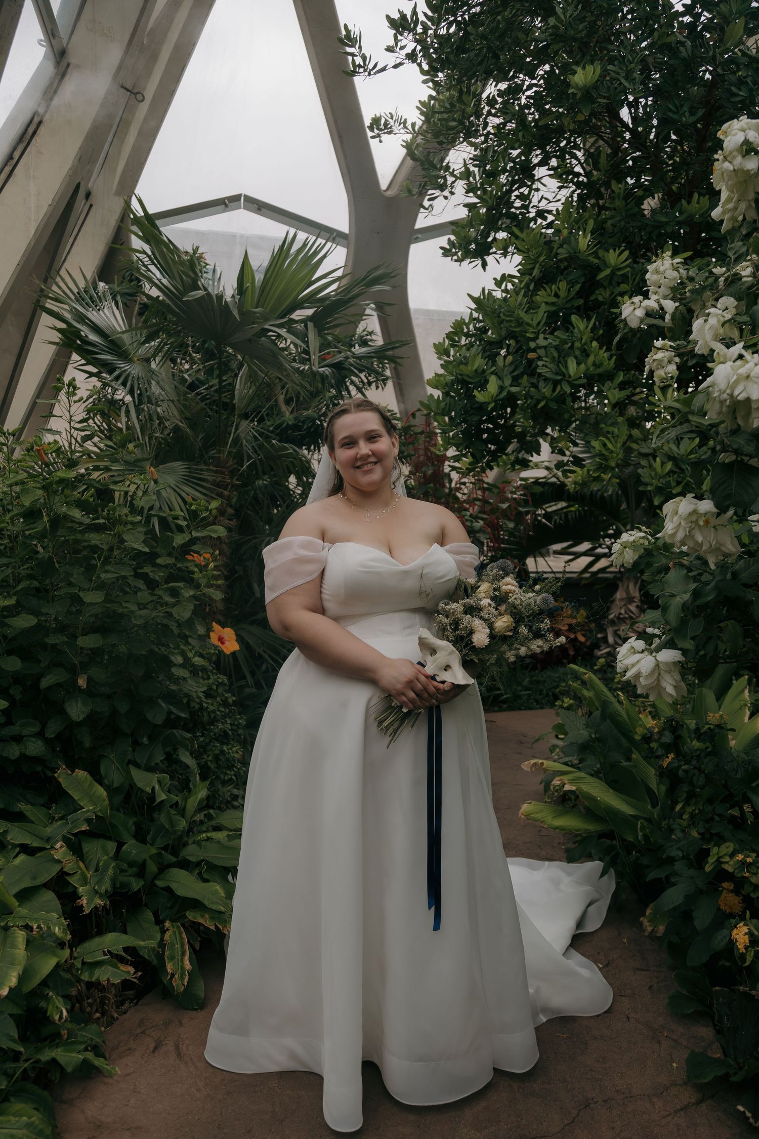 Smiling bride in an off-shoulder white ball gown holding a bouquet inside a lush botanical garden greenhouse.
