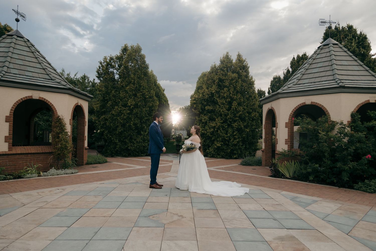 Bride and groom stand facing each other in a elegant courtyard garden at golden hour sunset.