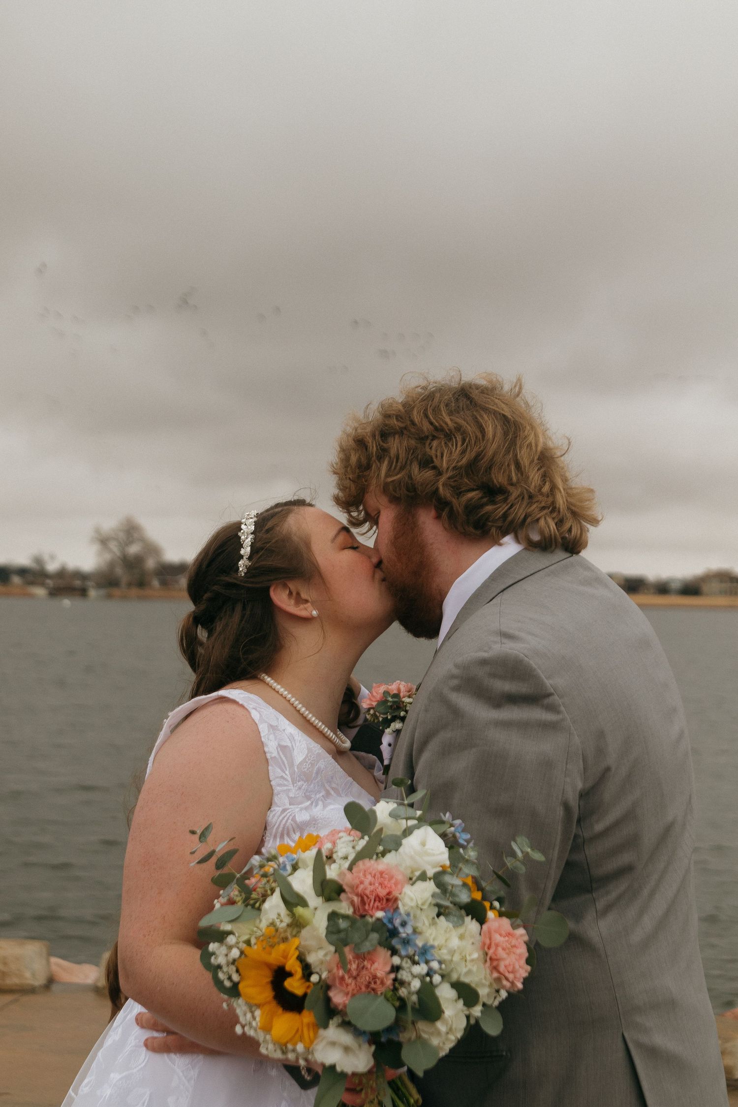 A couple shares a romantic moment against a cloudy sky while holding a vibrant bouquet with sunflowers.