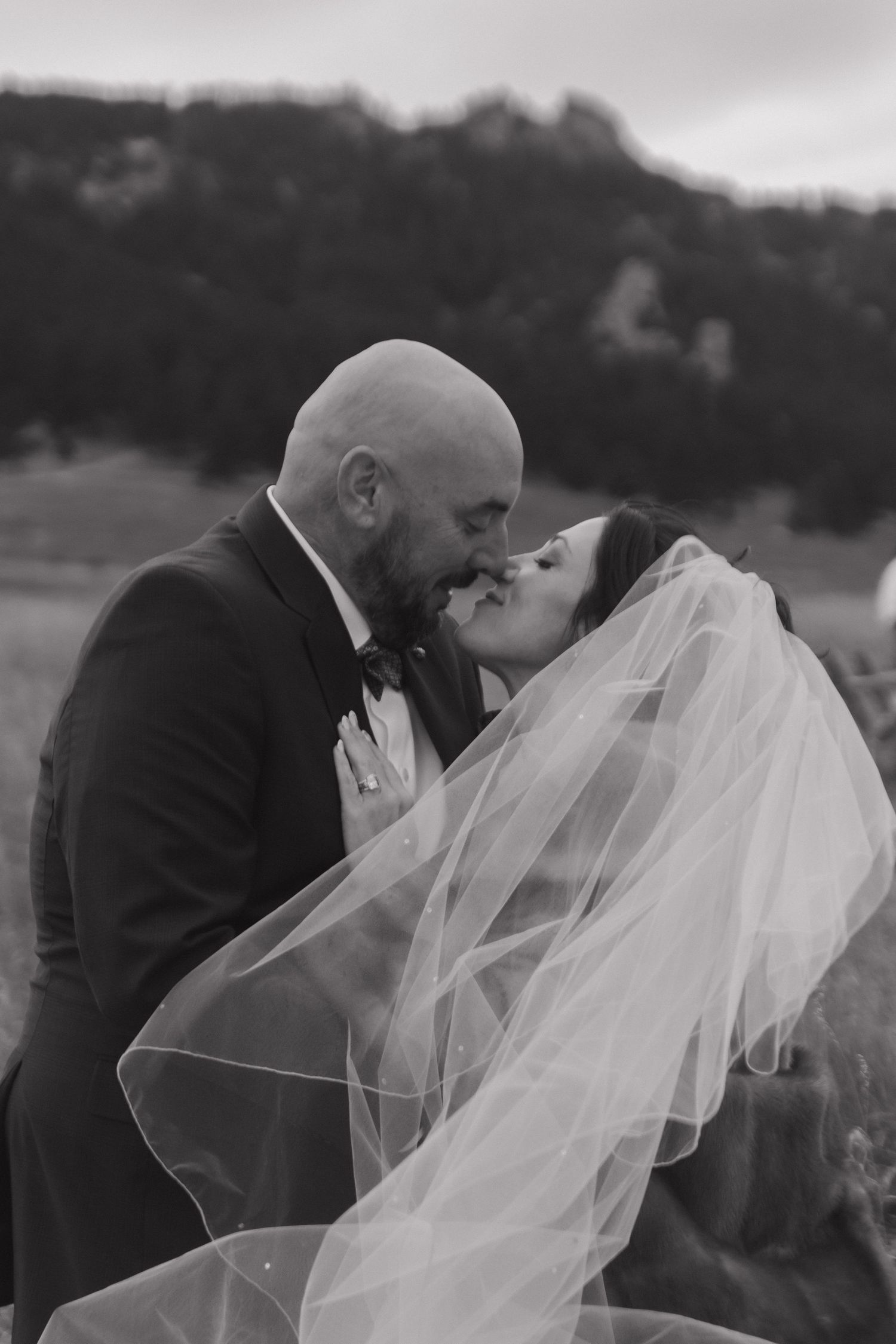 A romantic black and white wedding photo shows a couple sharing a tender kiss outdoors with a flowing veil in the breeze.