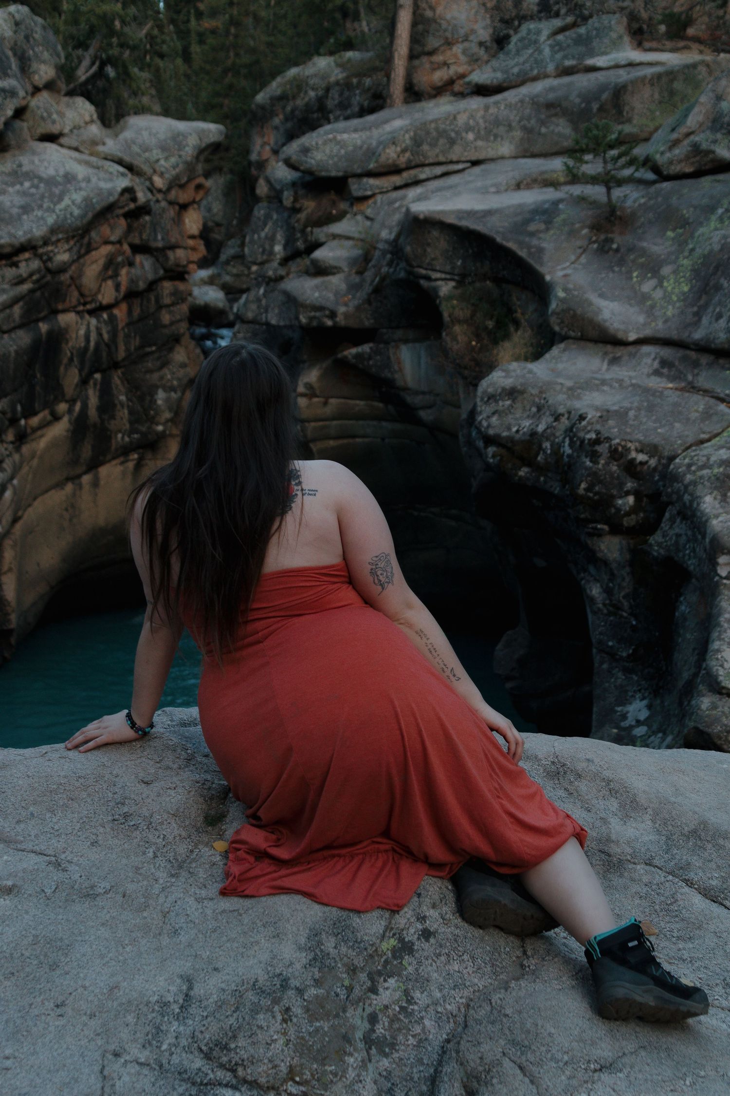 Person in red dress sits contemplatively on rocky cliffs overlooking serene blue waters in moody natural setting.