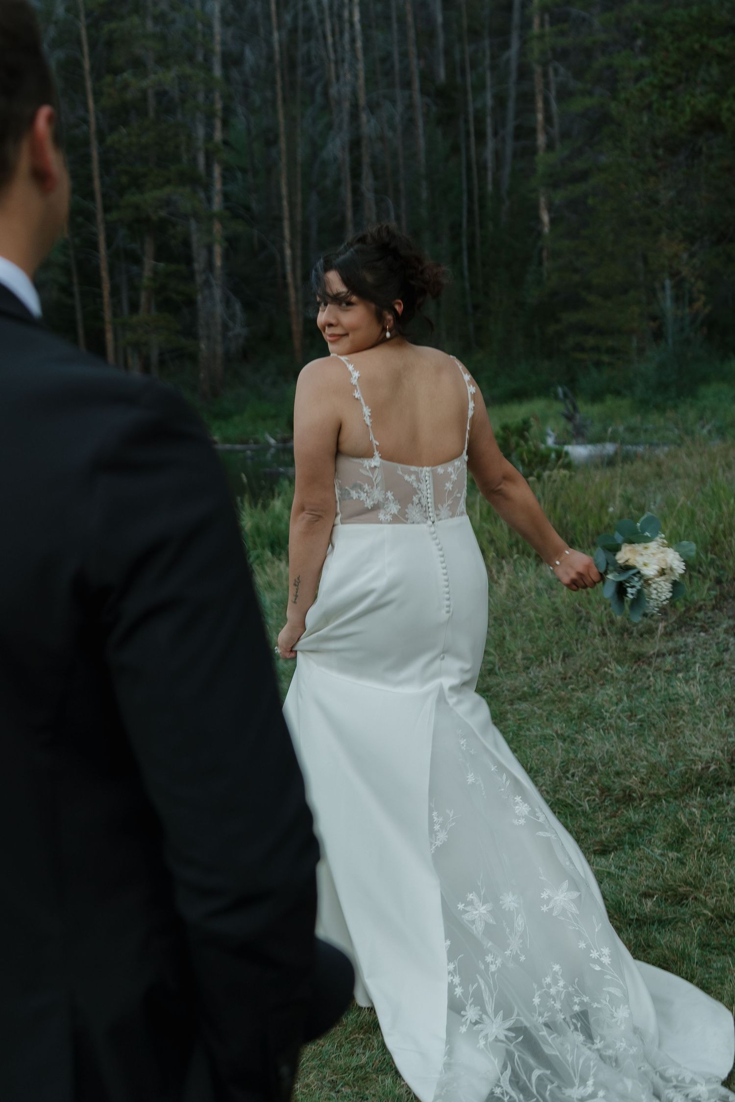 Bride in lace-detailed wedding gown walks along grassy lakeside path.