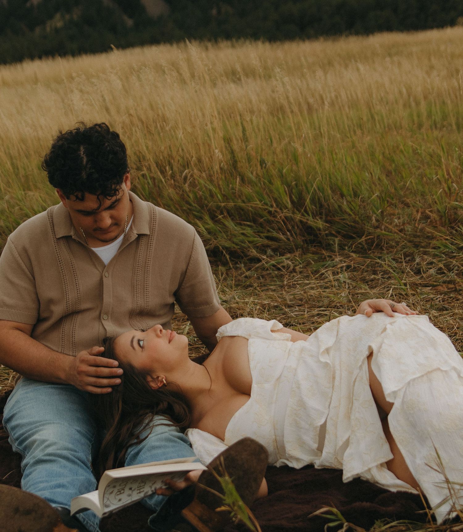 A romantic couple shares tender moments while sitting in a golden meadow with mountains behind them.