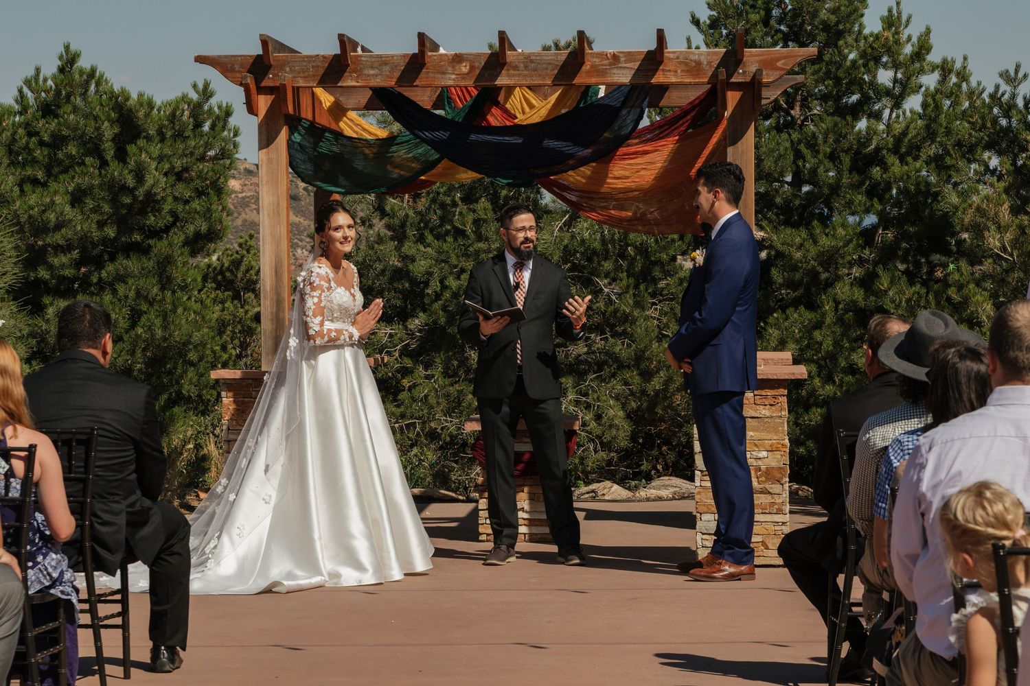 Wedding ceremony taking place under a rustic wooden pergola with draped fabric decor.