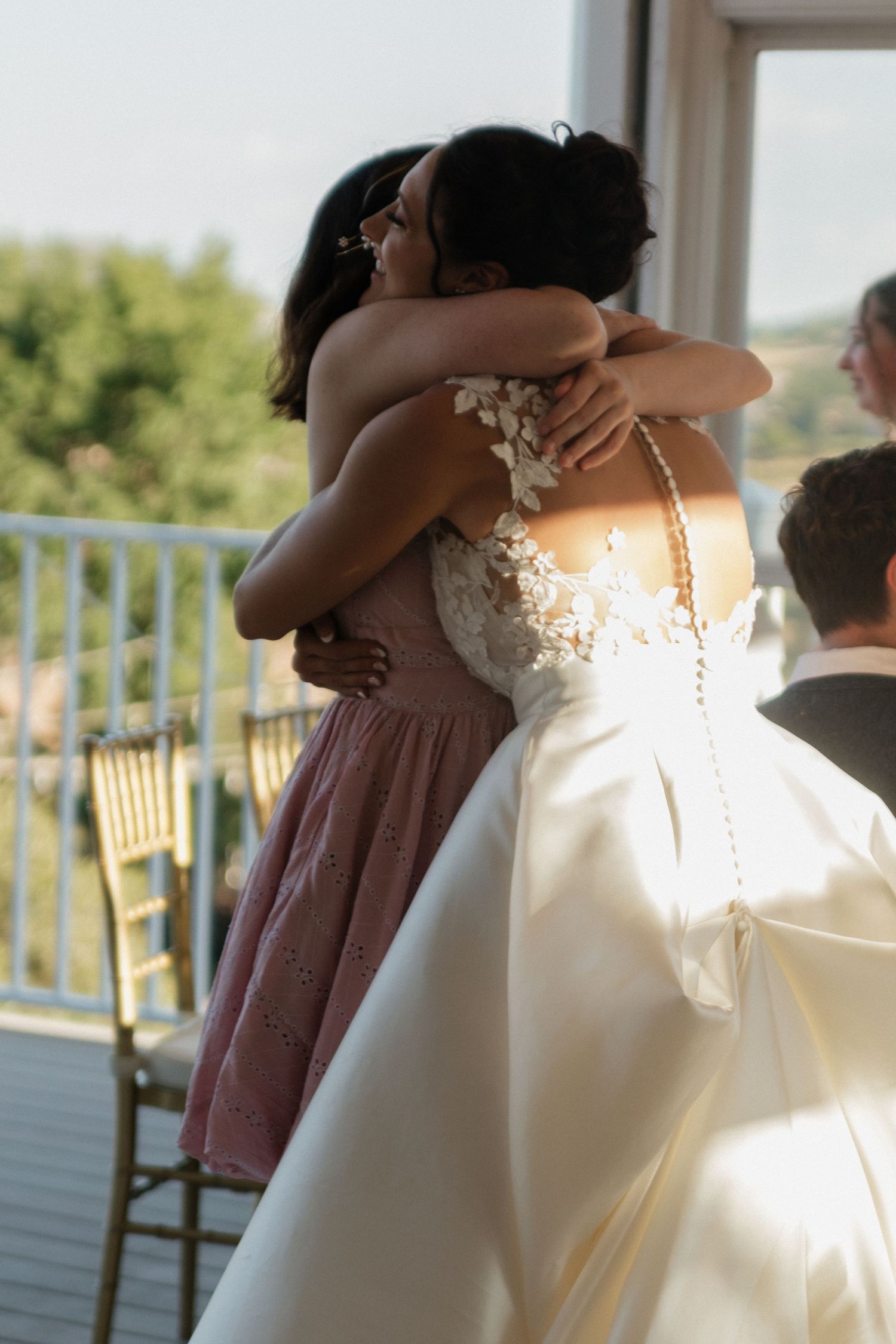 Wedding party members embrace each other emotionally on a sunlit balcony.
