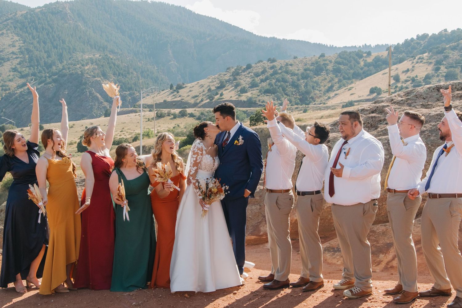 Wedding party in colorful attire poses in a line against mountain backdrop.
