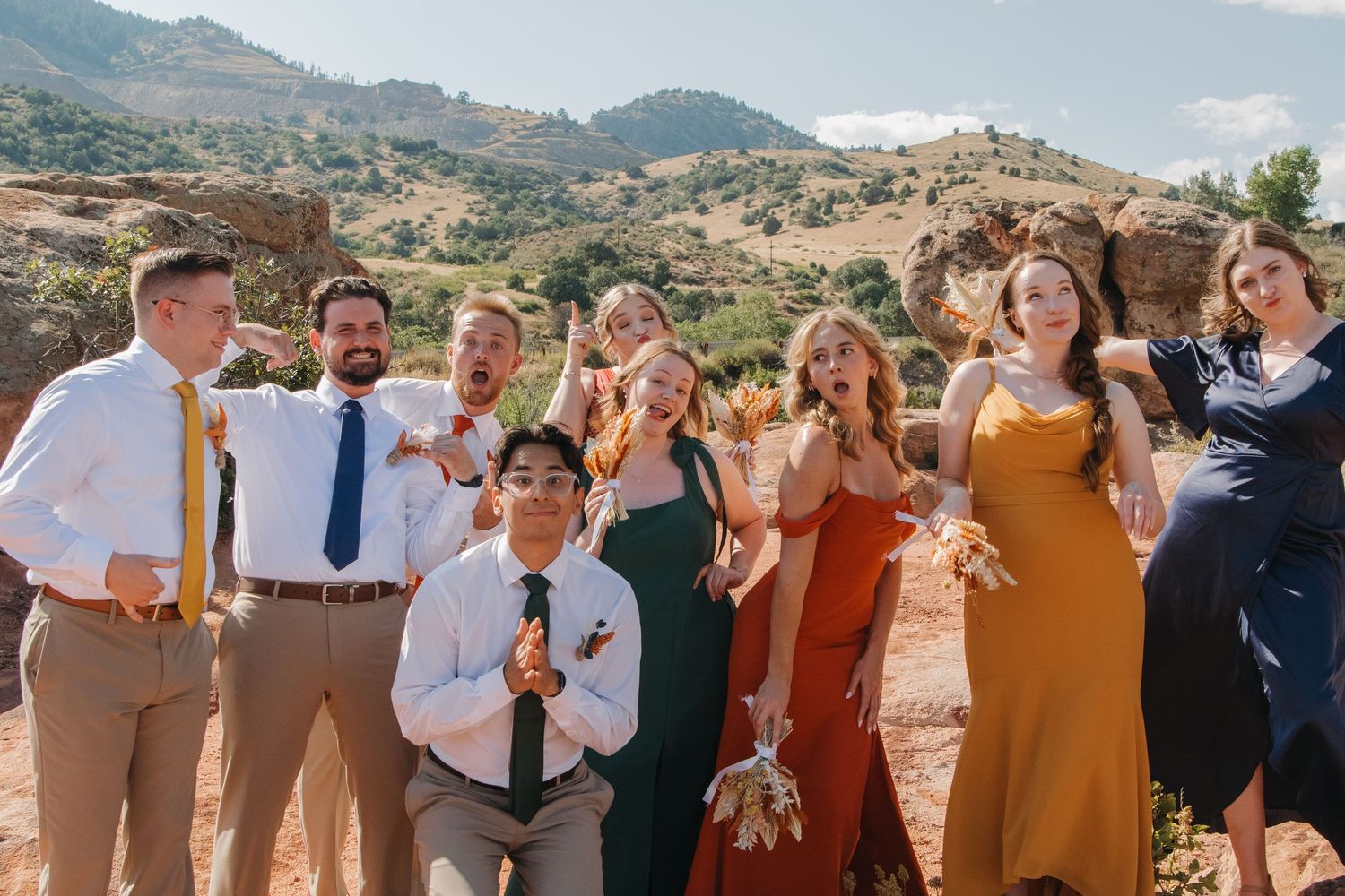 Wedding party members in autumn-toned attire pose together against mountain scenery.