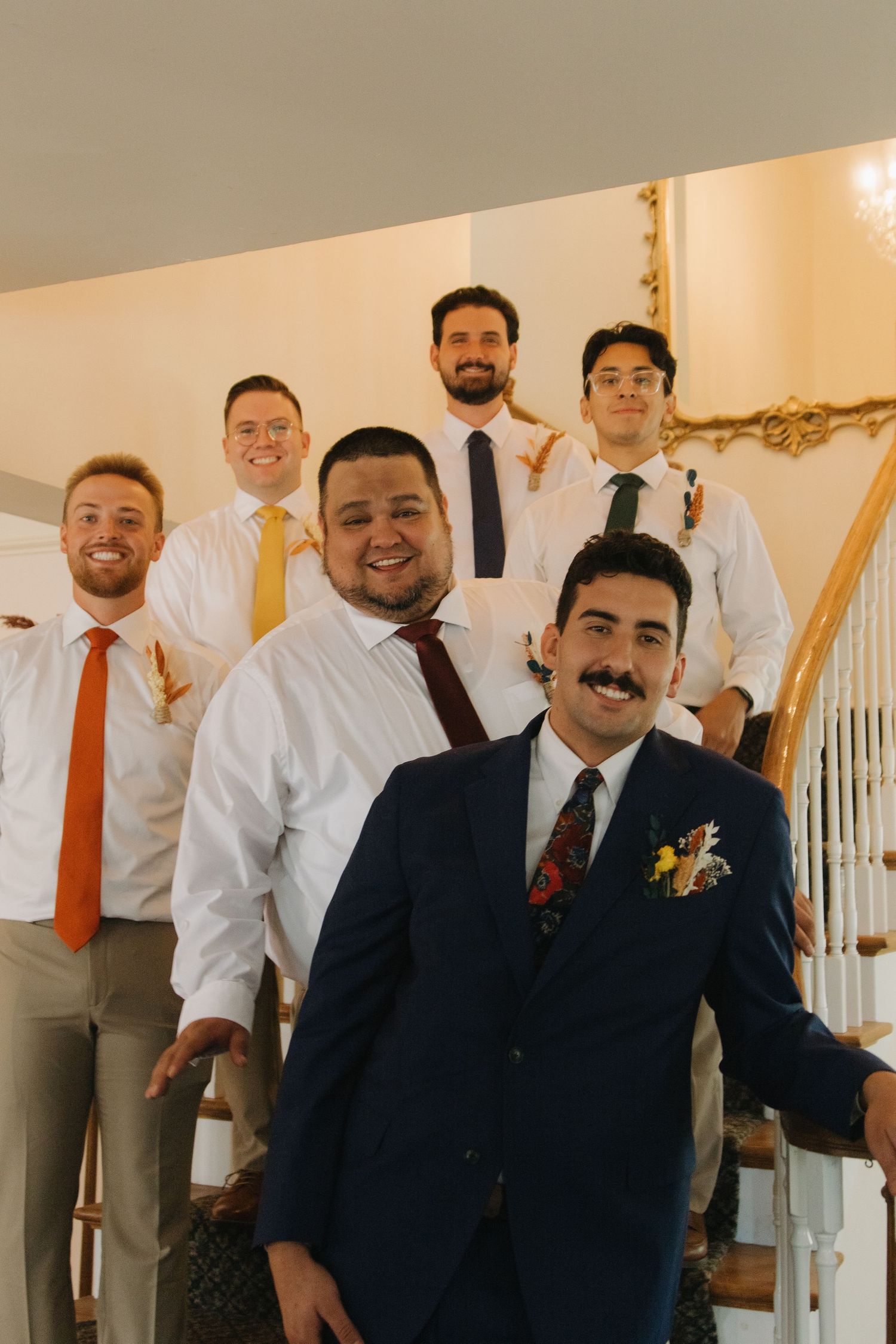 Wedding party members in formal attire gather at the bottom of an elegant staircase.