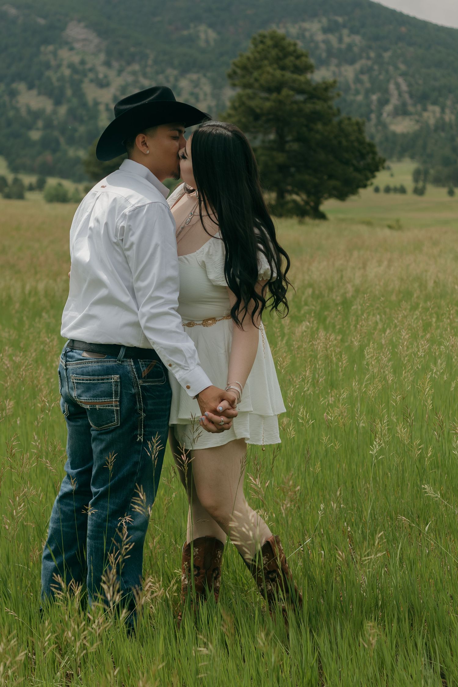 Couple in cowboy attire sharing an intimate moment in a grassy field with mountains in background.