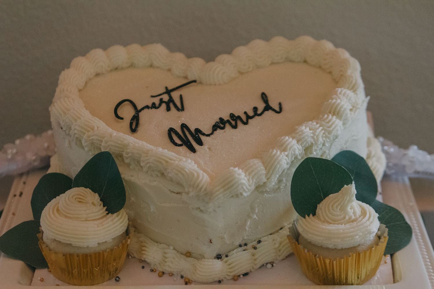 White wedding cake and cupcakes decorated with frosting details and writing on display.