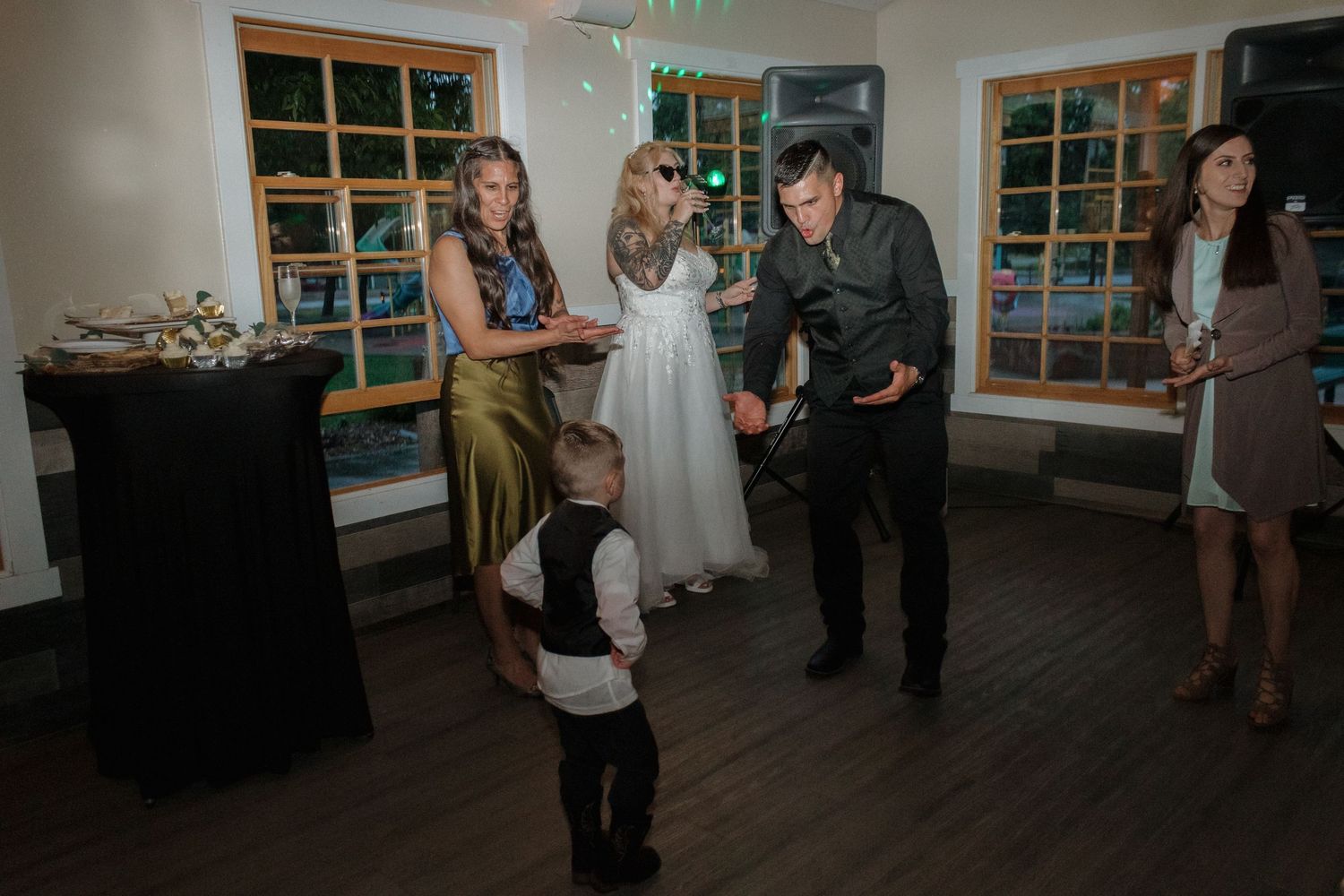 Wedding guests gather around a young ring bearer on a wooden dance floor.